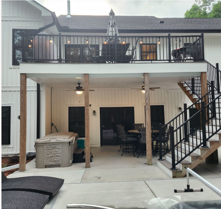 An image of the exterior of a residential home with a covered patio and balcony. The home has a gray exterior and a pitched roof. The patio has a metal railing and there are chairs and tables set up on the patio. The balcony above the patio has a metal railing as well. There is a staircase leading up to the balcony from the patio.