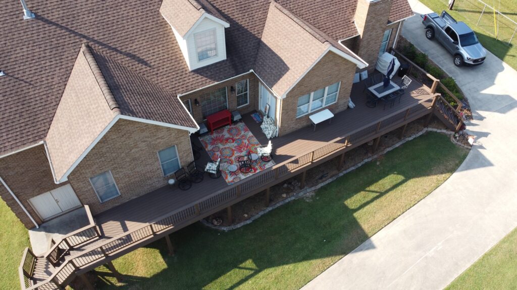 An aerial view of a large two-story house with a large wrap-around wooden deck and patio area, with a car parked in the driveway.