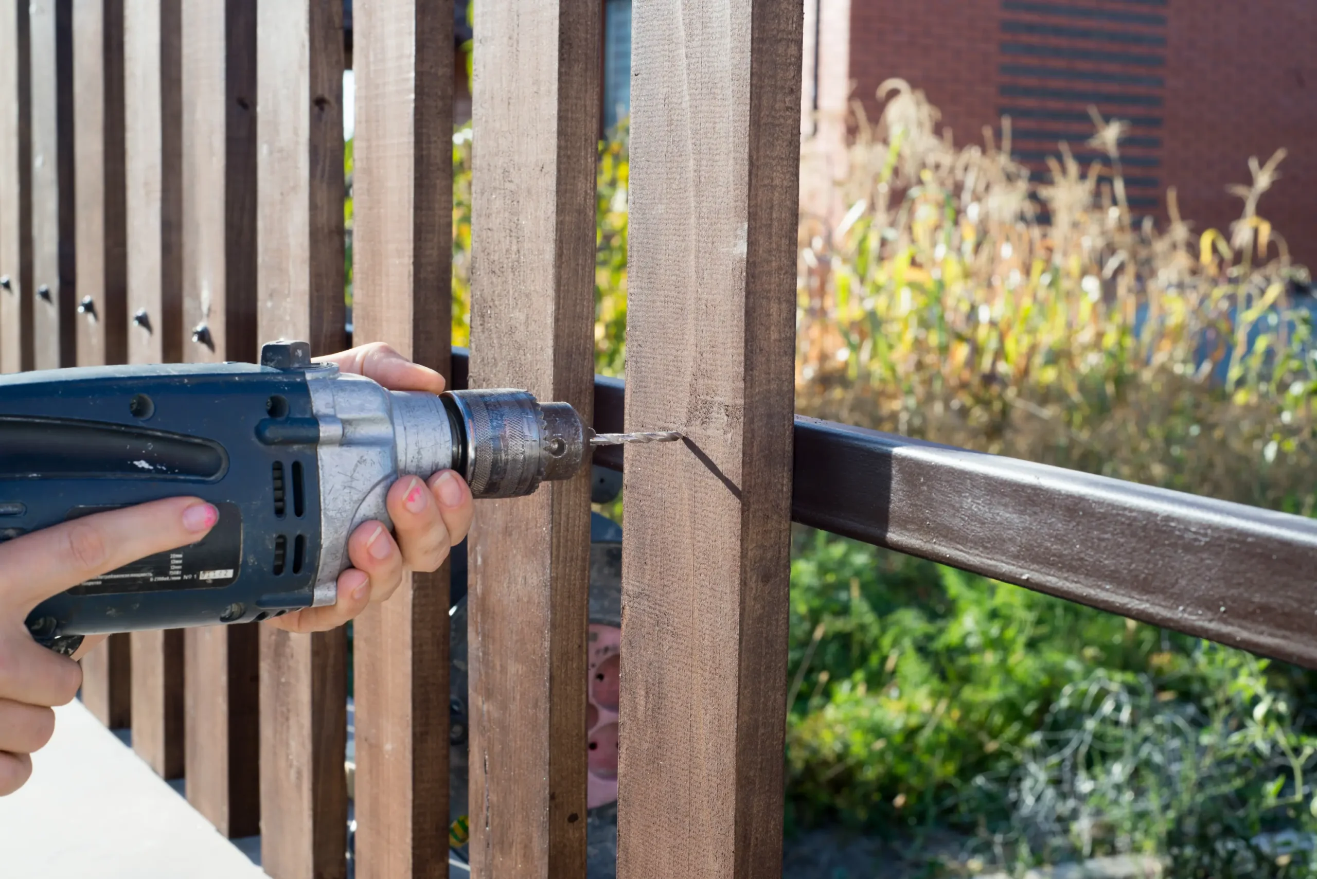 A power drill is being used to install or repair a wooden fence, with a lush green yard visible in the background.