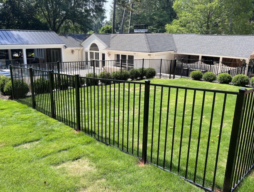 A black metal fence encloses a grassy area with a multi-building residential structure in the background, surrounded by trees.