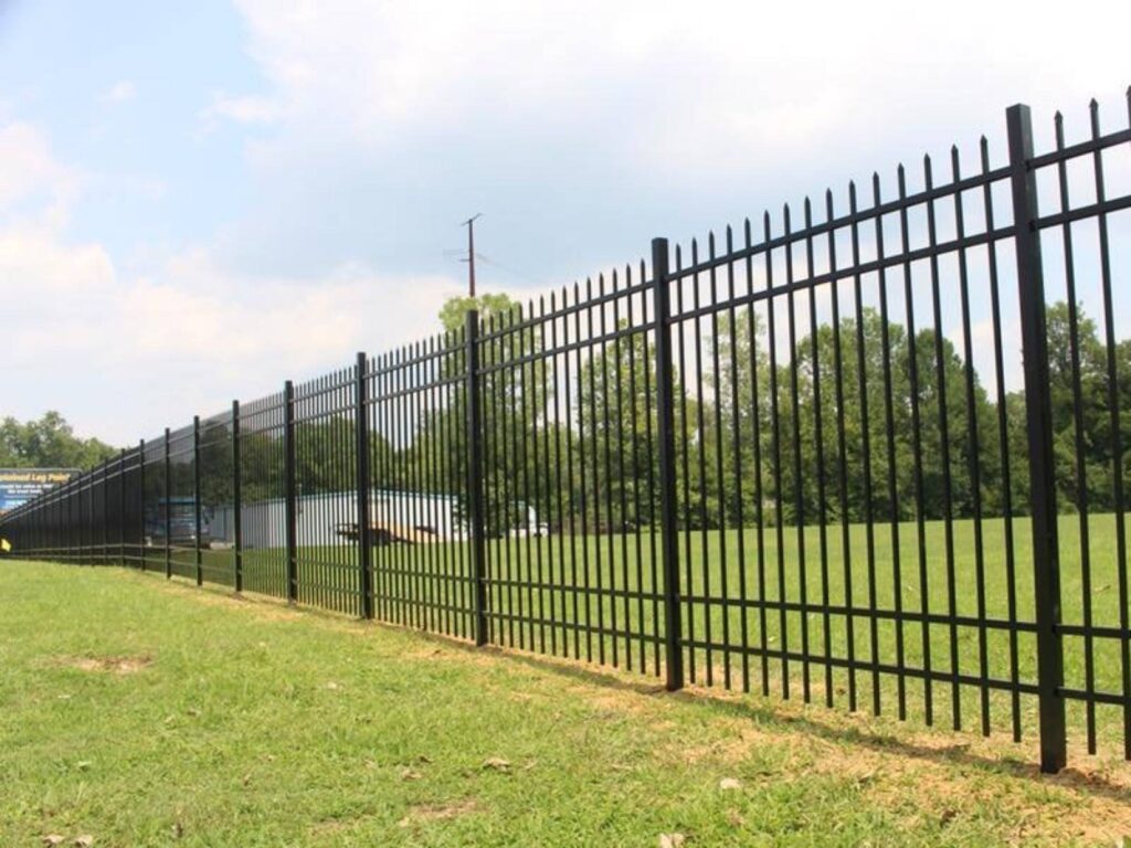 A long stretch of black metal fencing surrounding a grassy field, with trees and power lines visible in the background.