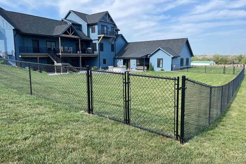 A two-story house with a large front porch and balcony, surrounded by a black metal chain-link fence and a grassy lawn.