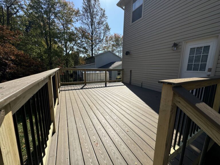 The image shows a wooden deck with a railing. The deck leads to a staircase that goes down to a grassy area. The house has a gray exterior with white trim and there are trees visible in the background.