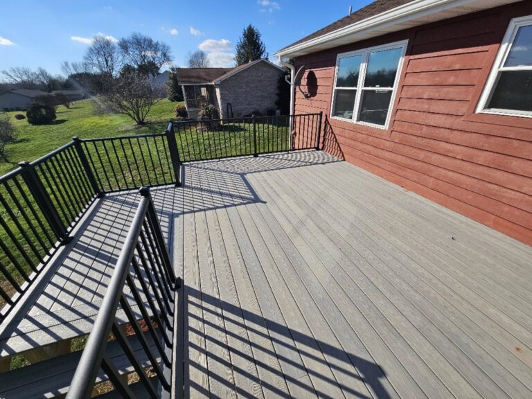 The image shows a large wooden deck with a railing. The deck extends out from a residential building with a red and brown exterior. There are trees and a grassy area visible in the background.