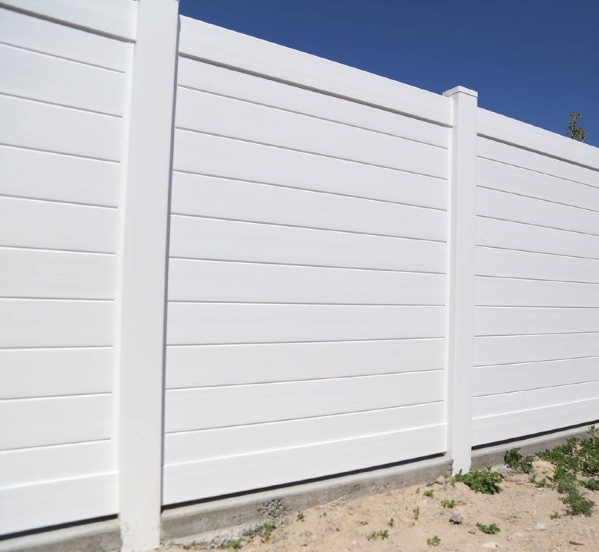 A tall white privacy fence with horizontal slats, standing in a grassy yard with trees in the background.