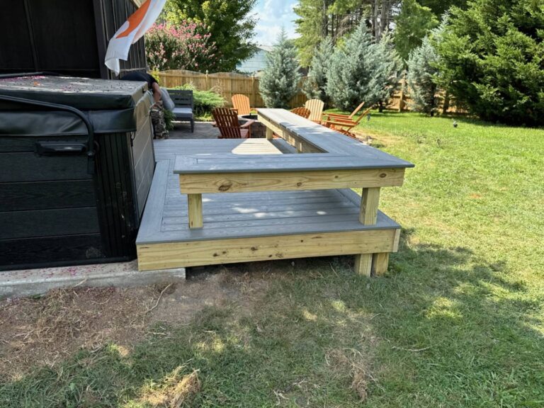 A wooden deck with stairs leading up to it, surrounded by trees and a grassy yard. A hot tub is visible in the background.