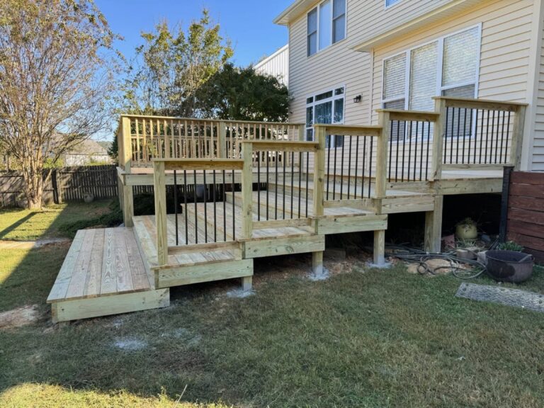 The image shows a large wooden deck and stairs leading up to a yellow house. The deck has a wooden railing and appears to have multiple levels, with a grassy yard in the foreground.