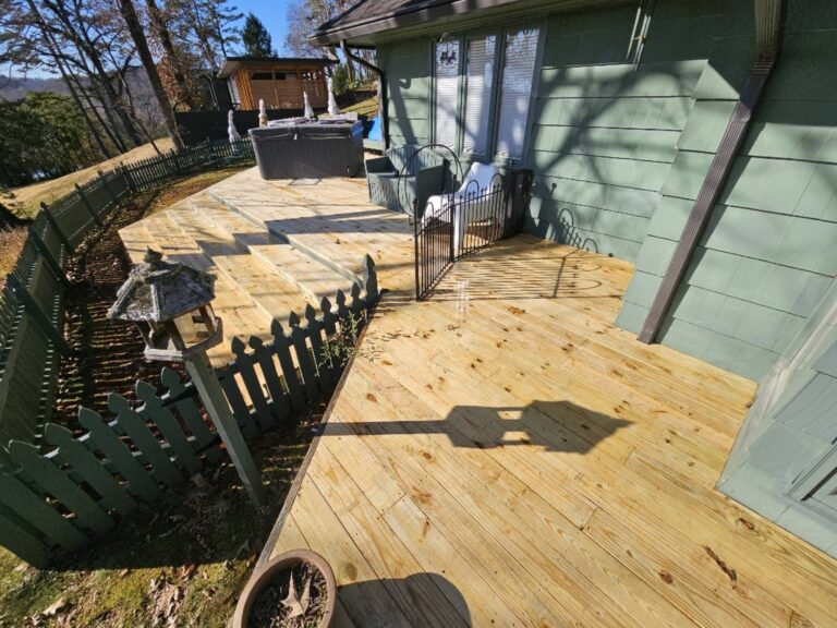 The image shows a wooden deck and stairs leading up to a green house. There is a grill and other outdoor equipment on the deck, as well as a wooden fence surrounding the property.