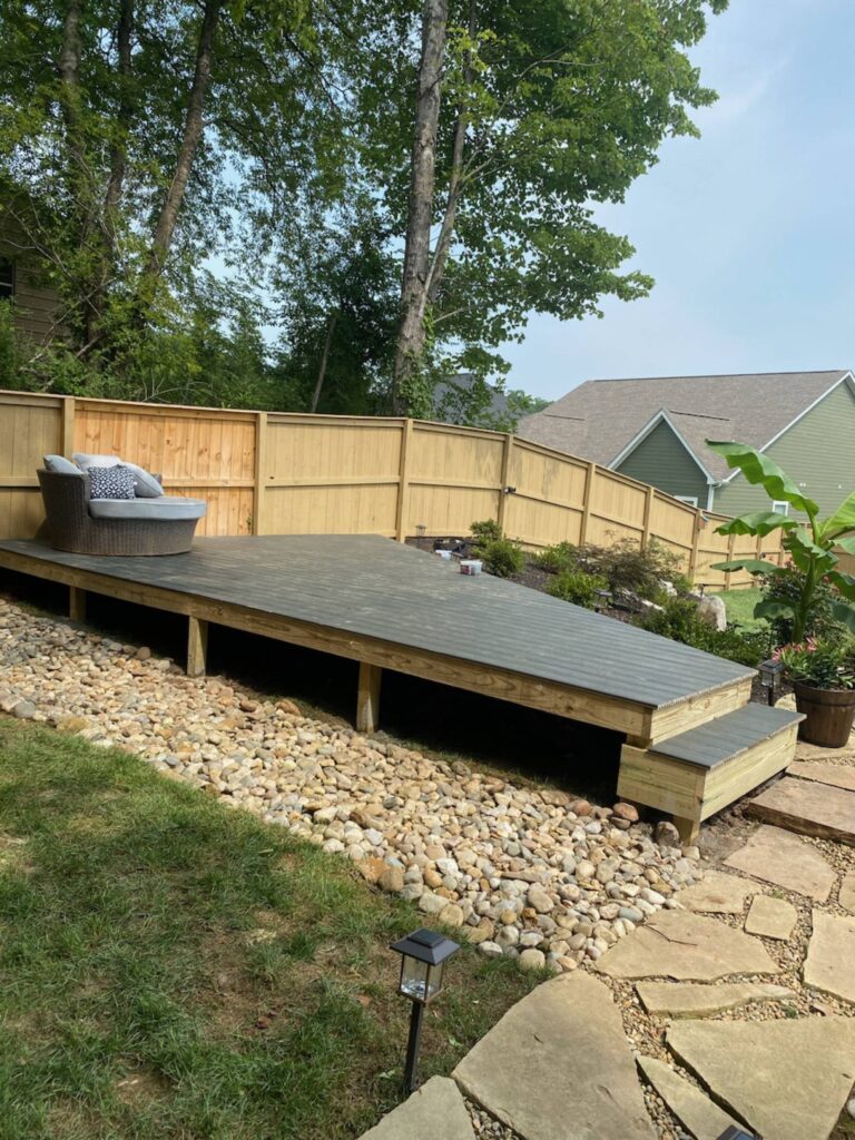 A large wooden deck with steps leading down to a grassy yard. The deck has a woven patio chair and several potted plants.