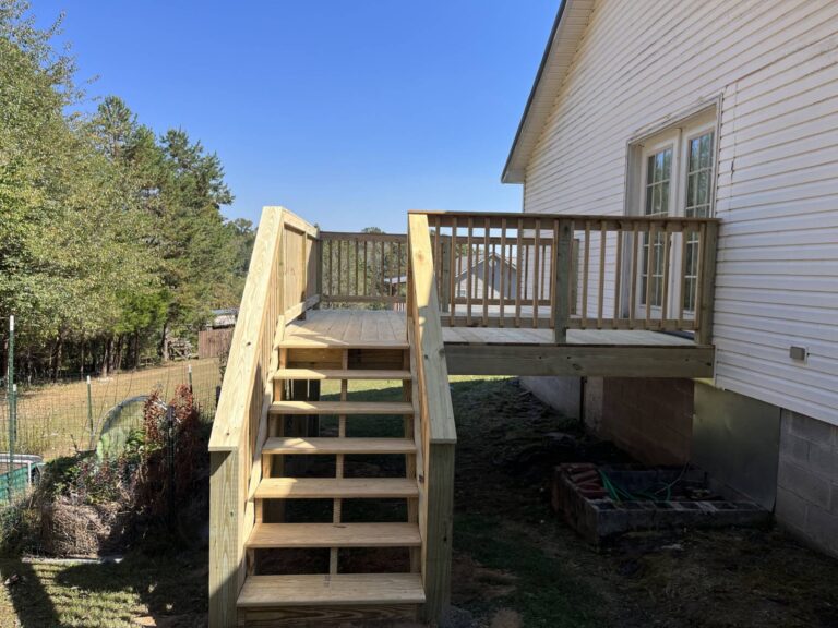 A newly constructed wooden deck and stairs leading up to a residential home, surrounded by trees and a fenced yard.