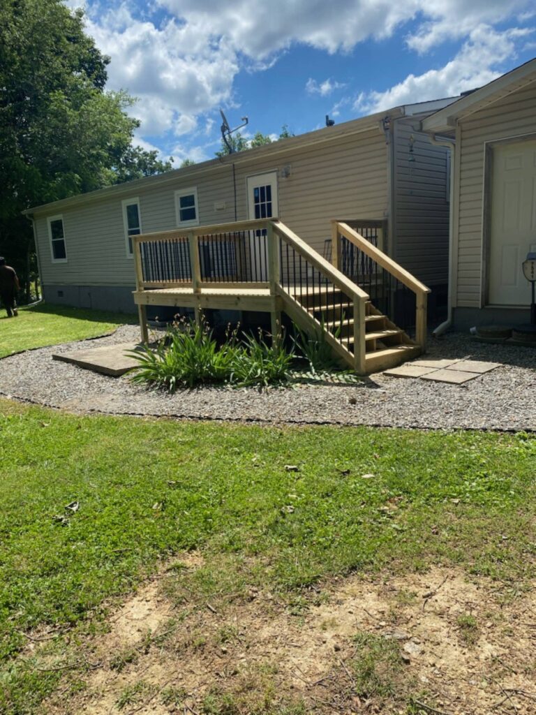 A wooden deck with stairs and railings, surrounded by trees and a grassy lawn.