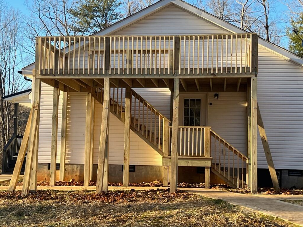 A wooden house with a large covered porch and wooden steps leading up to it, surrounded by trees.